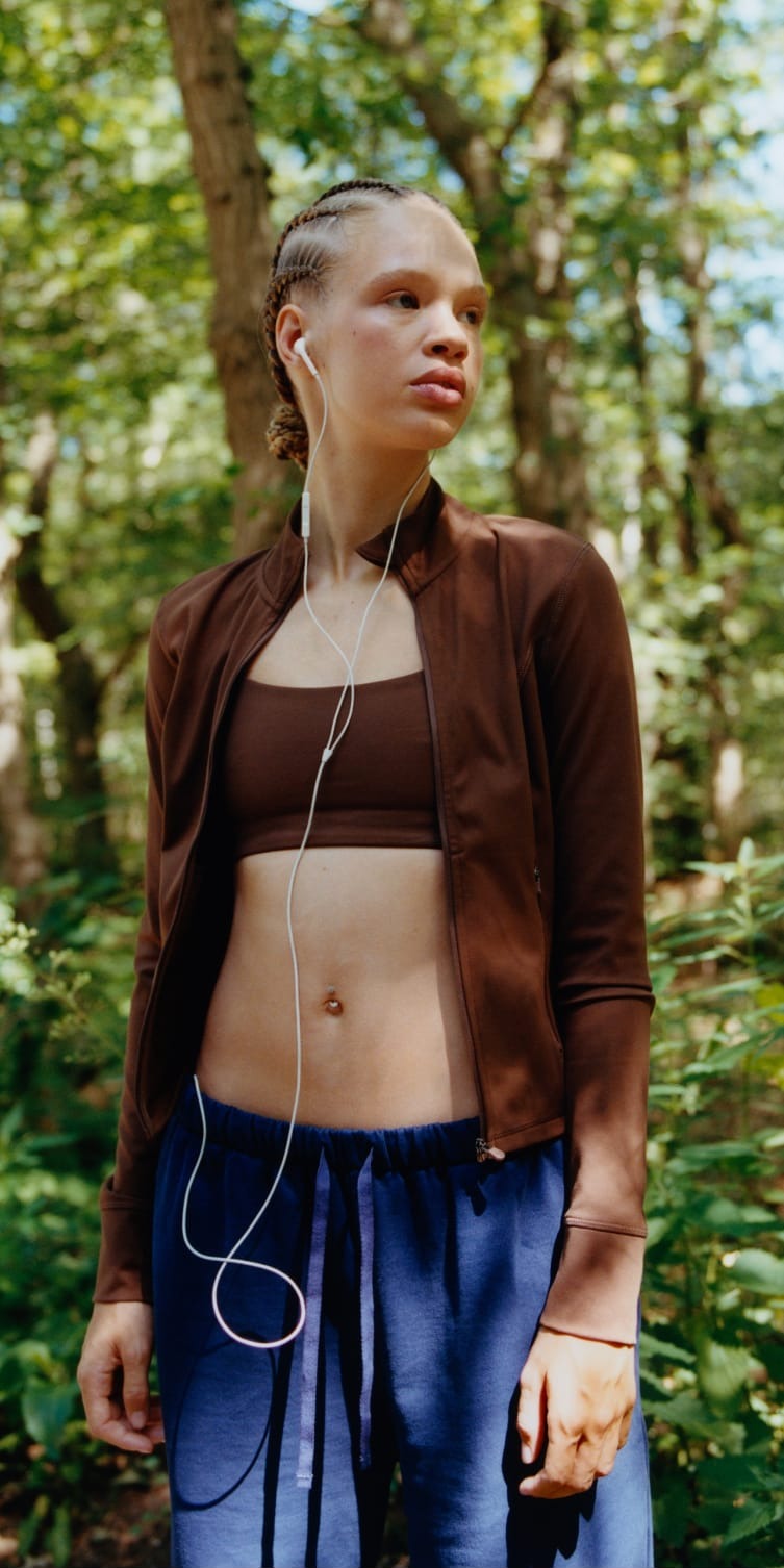 Two women outdoors in a sunlit forest wearing Organic Basics activewear: one stretching in a burgundy zip jacket and leggings, the other in a brown zip jacket and matching sports bra with wired earphones.